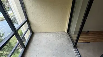 A long, narrow balcony features a concrete floor, a glass railing overlooking water and trees, and a sliding door leading to an Photo