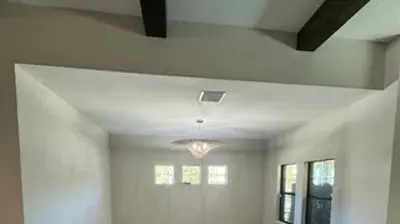 An empty, light gray room under construction, featuring unfinished concrete floors, a chandelier, and multiple windows. Photo