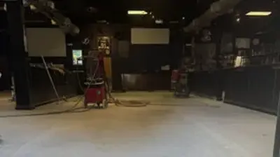 A wide shot of an empty room with a light concrete floor and dark walls, likely under renovation. Photo