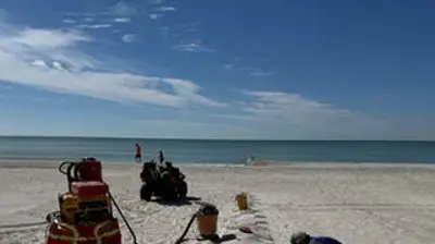 A person in a blue shirt installs a gray, segmented mat on a white sandy beach with the ocean and sky in the background. Photo