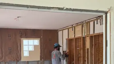 A construction worker performs selective demolition, revealing wall studs and insulation in a room. Photo