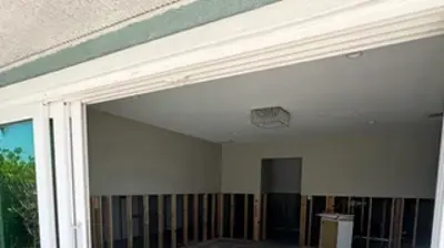 A room under renovation, viewed through open sliding glass doors, reveals exposed wall studs and a concrete floor. Photo