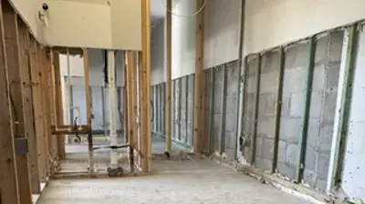 An interior room under construction shows exposed wooden wall studs and concrete block walls, with a bare concrete floor. Photo