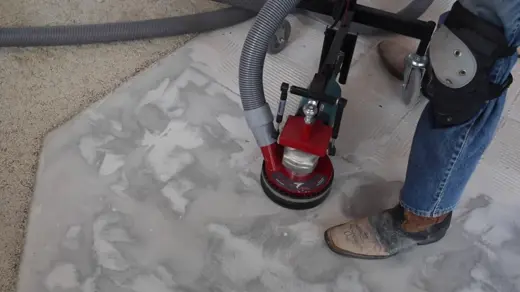 A person operates a dust-extracting floor grinder on a light-colored, patterned surface, wearing blue jeans and knee pads.