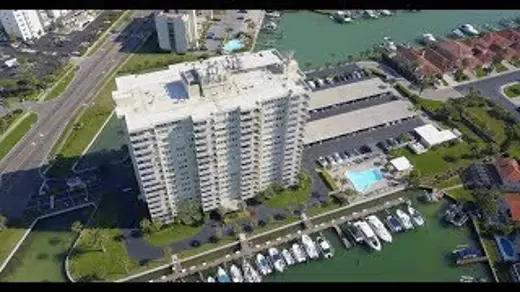 Aerial view of a tall, modern building situated beside a marina filled with boats, featuring a swimming pool and a road.