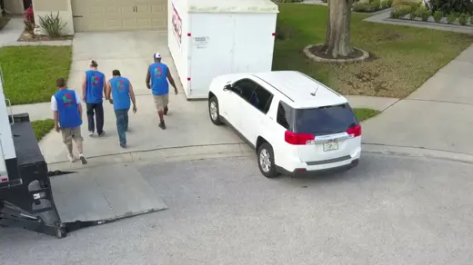 Movers load a moving truck and a large storage container on a residential driveway.