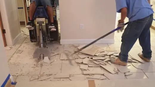 Two workers remove floor tiles; one operates a large machine, while the other scrapes debris.
